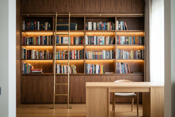 Floor-to-ceiling walnut library wall with lit shelves, brass ladder and a white oak desk